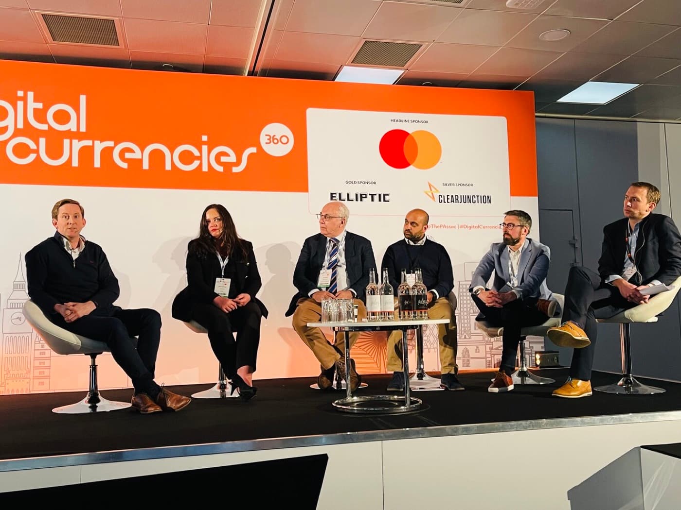 Panel of six speakers seated on stage at the Digital Currencies 360 conference in London, with Mastercard as headline sponsor.